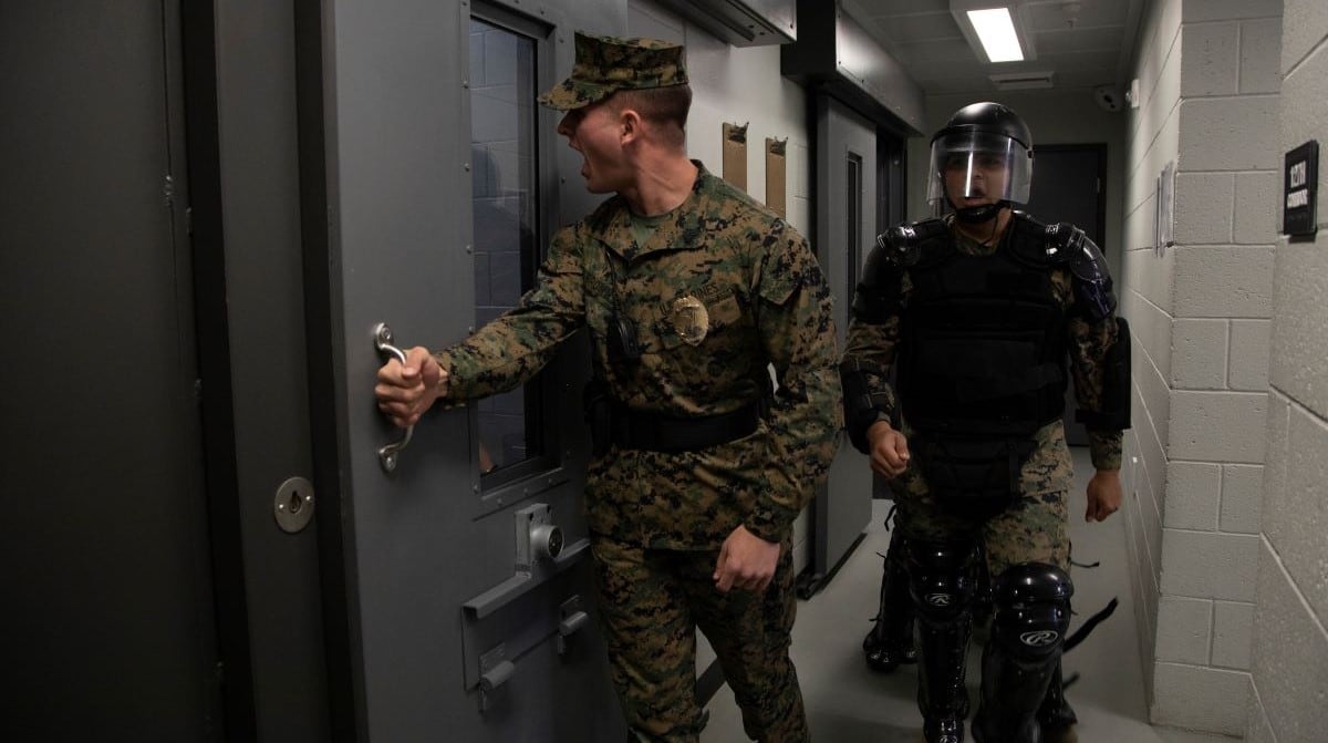 A male Marine in camis in the center opens the door to a brig cell. Another male Marine in protective gear trails behind him.